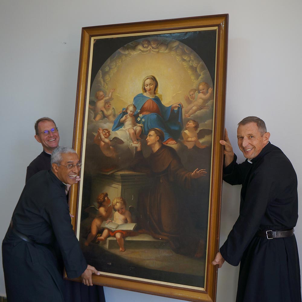 Três cônegos seguram um quadro de Nossa Senhora com Santo Antônio e anjos.